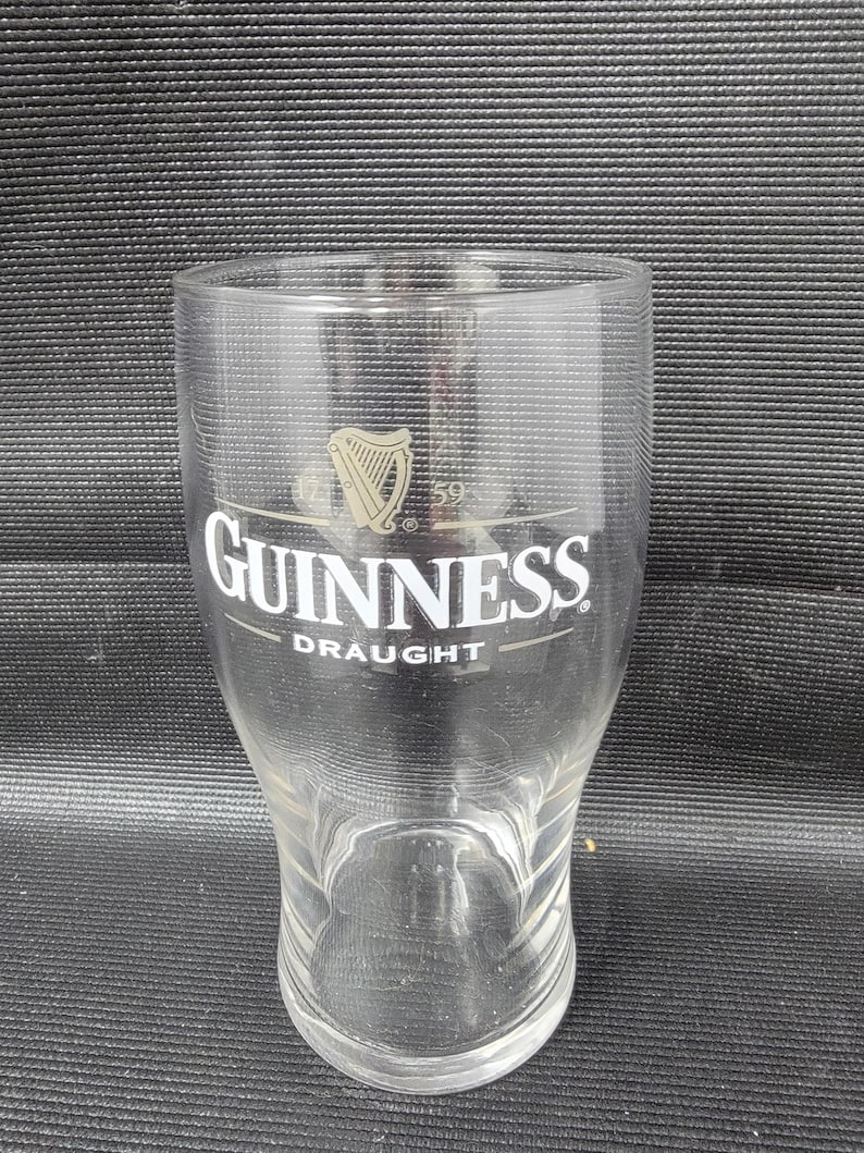 May include: A clear pint glass with the Guinness logo, featuring a harp and the word "Guinness" in white lettering. The word "Draught" is printed below. The glass has a slightly curved shape and a wide mouth.
