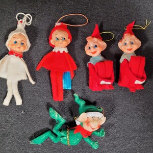 May include: Six vintage Christmas elf ornaments. The elves are made of felt and have painted faces. One elf is wearing a white suit, one is wearing a red suit, and four are wearing red and blue suits.