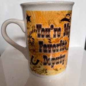 K&ouml;nnte beinhalten: Wei&szlig;er Keramikbecher mit gebogenem Henkel. Der Becher zeigt den Text "You're My Favorite Deputy" in Schwarz und Orange, zusammen mit einem Cowboyhut, Stern und Hufeisen-Design auf orangefarbenem Hintergrund.