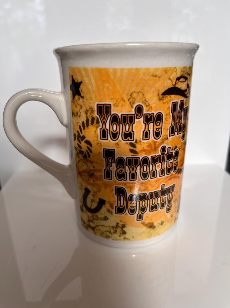 K&ouml;nnte beinhalten: Wei&szlig;er Keramikbecher mit gebogenem Henkel. Der Becher zeigt den Text "You're My Favorite Deputy" in Schwarz und Orange, zusammen mit einem Cowboyhut, Stern und Hufeisen-Design auf orangefarbenem Hintergrund.