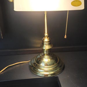 May include: A brass-colored desk lamp with a white, curved shade. The lamp has a round base, a slender stem, and a pull chain. A small sticker is visible on the shade. The lamp is plugged into an electrical outlet.