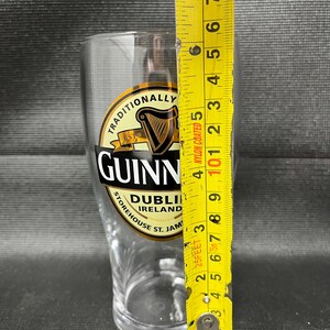 May include: A clear Guinness pint glass with the Guinness logo and text. The glass is measured with a yellow tape measure, showing a height of approximately 6 inches.