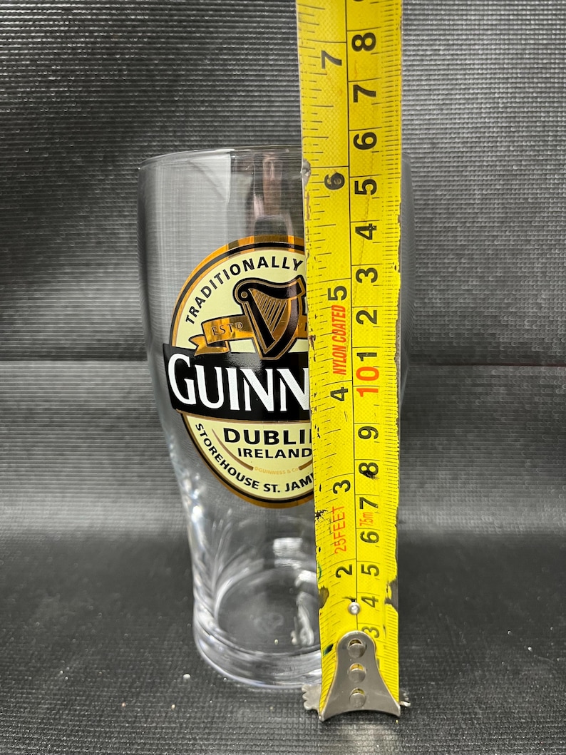 May include: A clear Guinness pint glass with the Guinness logo and text. The glass is measured with a yellow tape measure, showing a height of approximately 6 inches.