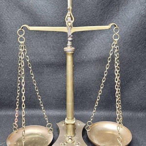 May include: A vintage brass balance scale with two round pans hanging from chains. The scale has a decorative base and a tall, slender column.