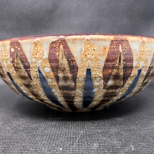 May include: A handcrafted ceramic bowl with a distinctive design. The bowl has a cream-coloured base with brown and blue vertical patterns. The rim has a textured appearance, and the bowl is suitable for serving food or as a decorative item.