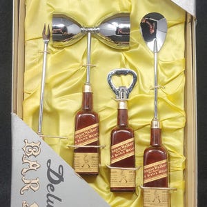 May include: A set of bar tools in a box with a yellow satin lining. The tools include a cocktail shaker, a bottle opener, a jigger, and a cocktail fork. The box is labeled "Deluxe Bar Set" and "Triple Plated Chrome".