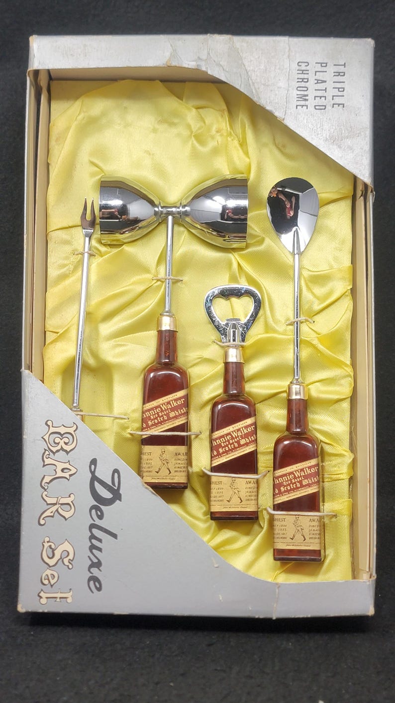 May include: A set of bar tools in a box with a yellow satin lining. The tools include a cocktail shaker, a bottle opener, a jigger, and a cocktail fork. The box is labeled "Deluxe Bar Set" and "Triple Plated Chrome".