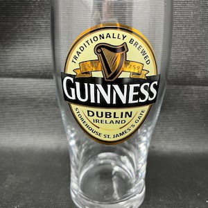 May include: A clear pint glass with the Guinness logo. The logo features a gold harp, the words "Guinness" in white, and the text "Dublin Ireland Storehouse St. James's Gate". The glass is designed for serving beer.