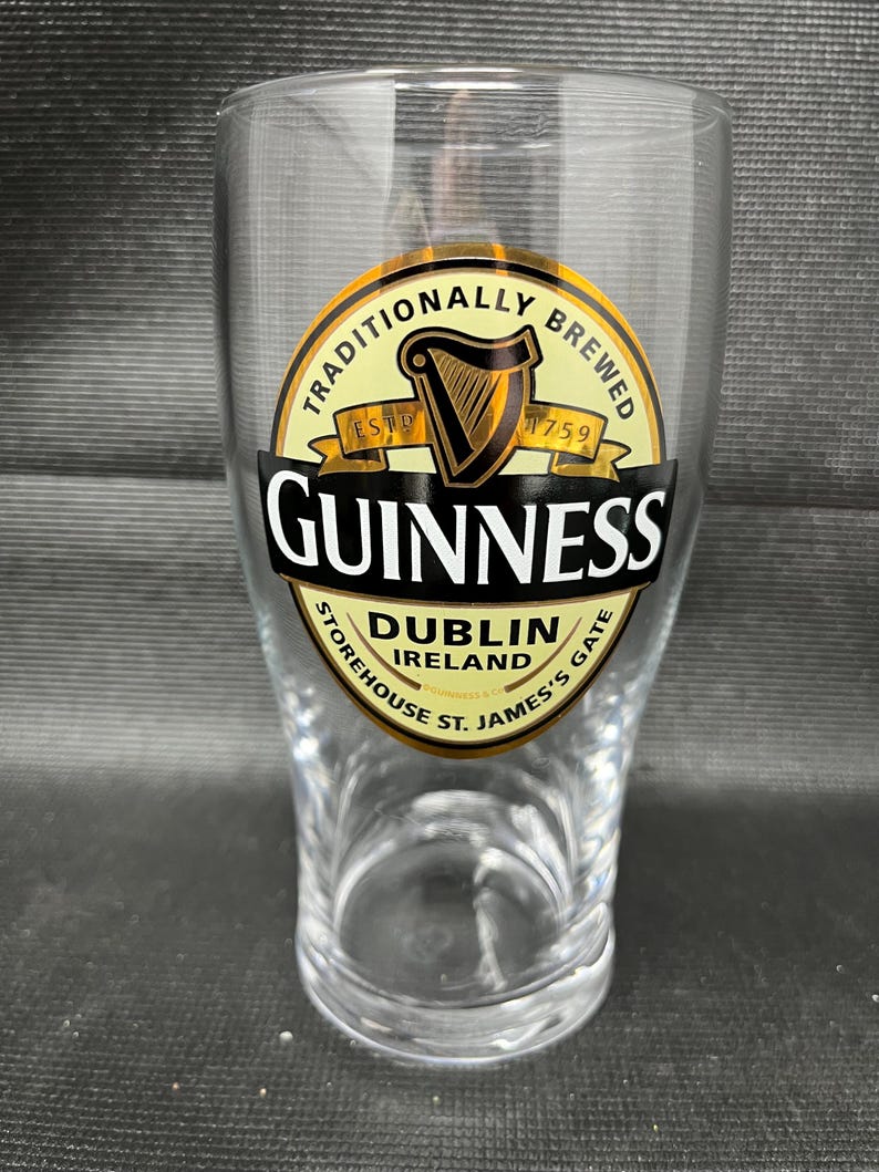 May include: A clear pint glass with the Guinness logo. The logo features a gold harp, the words "Guinness" in white, and the text "Dublin Ireland Storehouse St. James's Gate". The glass is designed for serving beer.