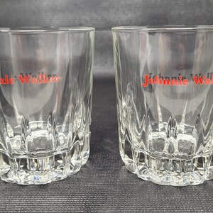 May include: Two clear, faceted whiskey glasses with the red "Johnnie Walker" logo printed on the front. The glasses have a thick, textured base and a slightly flared rim. The glasses are empty and sit on a dark surface.