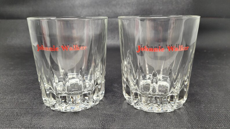 May include: Two clear, faceted whiskey glasses with the red "Johnnie Walker" logo printed on the front. The glasses have a thick, textured base and a slightly flared rim. The glasses are empty and sit on a dark surface.