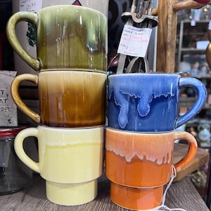 May include: A stack of four ceramic mugs featuring a glossy drip glaze finish. The mugs are coloured in olive green, brown, yellow, blue, and orange. Each mug has a side handle and a cylindrical body with a slightly recessed base for stacking.