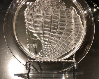 May include: A clear glass plate with a raised, detailed design of a seashell. The plate is on a stand and the year "1973" is etched on the bottom.
