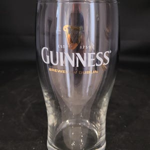 May include: A clear Guinness pint glass with the Guinness logo in white and gold. The glass has a curved shape and is empty. The text "EST 1759" and "BREWED IN DUBLIN" are also visible.