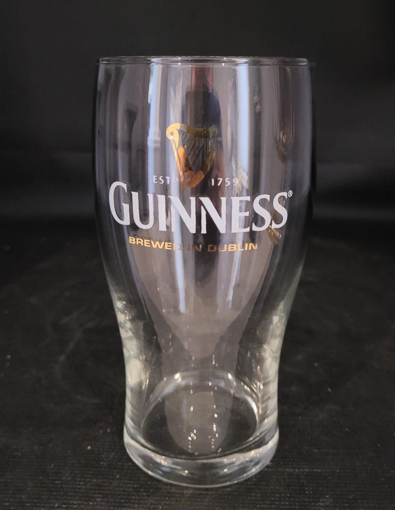 May include: A clear Guinness pint glass with the Guinness logo in white and gold. The glass has a curved shape and is empty. The text "EST 1759" and "BREWED IN DUBLIN" are also visible.