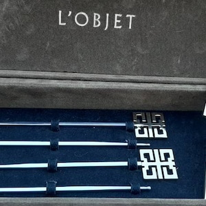 May include: A silver wine stopper and four silver cocktail picks with a geometric design, presented in a grey box. The box is lined with dark blue fabric and has the brand name "L'OBJET" printed on the inside of the lid.