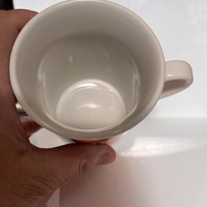 K&ouml;nnte beinhalten: Eine wei&szlig;e Keramik-Kaffeetasse mit einem abgerundeten Henkel. Das Innere der Tasse ist wei&szlig;, und die Au&szlig;enseite hat ein orangefarbenes Band in der N&auml;he des Bodens. Die Tasse wird von einer Hand gehalten.