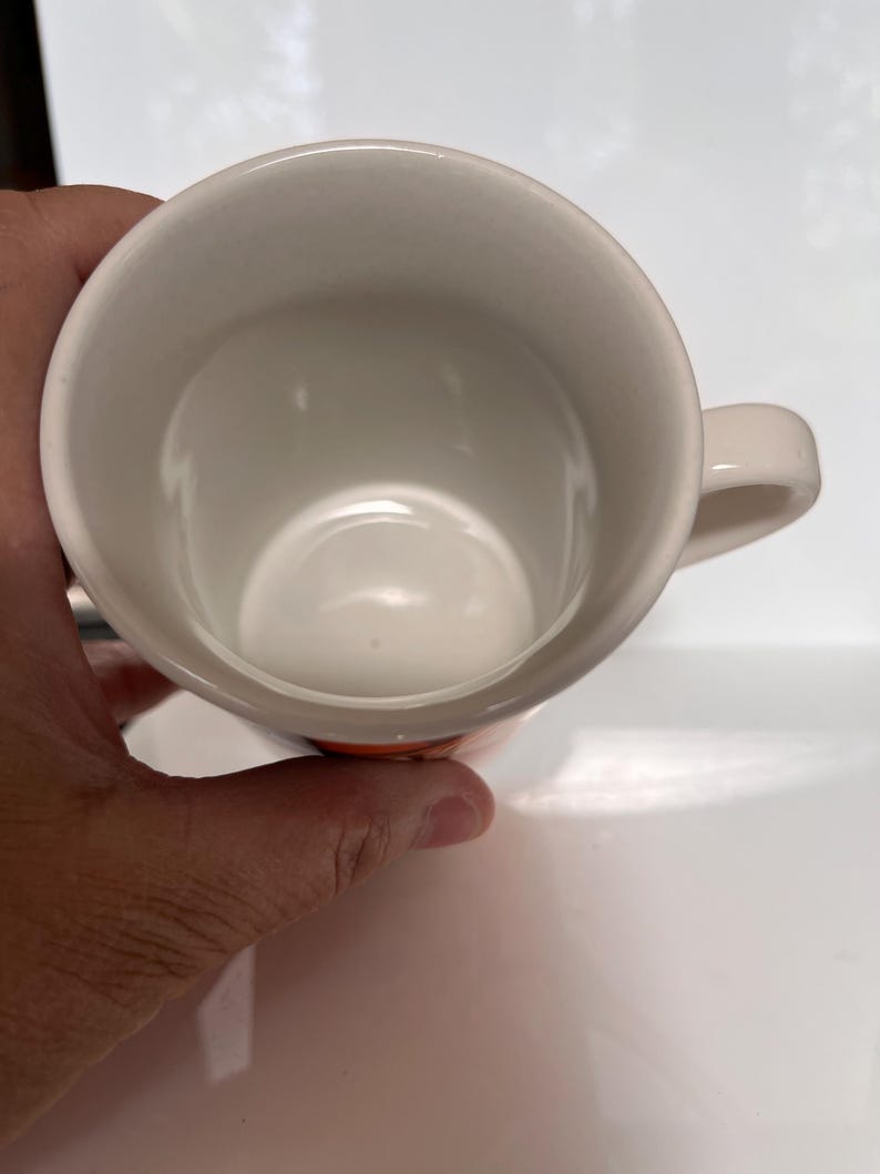 K&ouml;nnte beinhalten: Eine wei&szlig;e Keramik-Kaffeetasse mit einem abgerundeten Henkel. Das Innere der Tasse ist wei&szlig;, und die Au&szlig;enseite hat ein orangefarbenes Band in der N&auml;he des Bodens. Die Tasse wird von einer Hand gehalten.