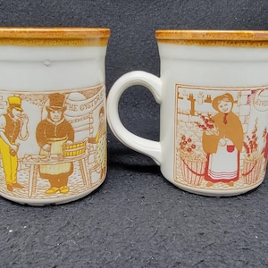 May include: Two white ceramic mugs with brown trim and illustrations. One mug features a scene of people selling oysters, the other features a woman selling flowers. Both mugs have the text "The Oysters" printed on them.