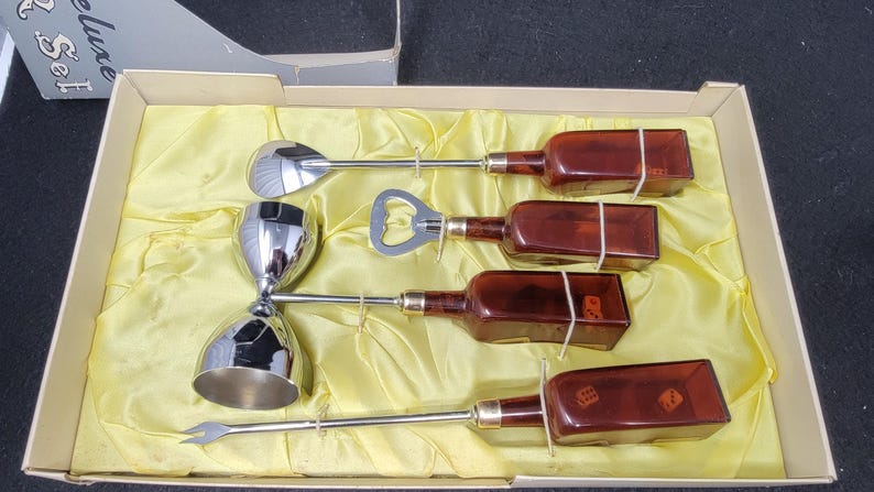 May include: A set of five silver cocktail tools in a yellow velvet-lined box. The set includes a jigger, a bottle opener, a spoon, and two small bottles with dice inside.