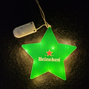 May include: A green star-shaped light with the Heineken logo in white and a red star. The light is connected to a small white battery pack by a thin wire. The star has a textured surface.