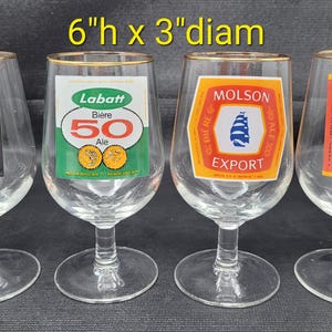 May include: Four clear beer glasses with gold rims, each with a different beer label. The labels include "Biere O'Keefe Ale", "Labatt 50 Ale", "Molson Export", and "Carling Black Label Biere Beer". The glasses are approximately 6 inches high and 3 inches in diameter.