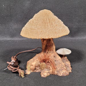 May include: A mushroom-shaped lamp with a textured, beige lampshade. The lamp's stem and base are brown, with a small mushroom and decorative foliage. The lamp has a brown cord and a two-prong plug.