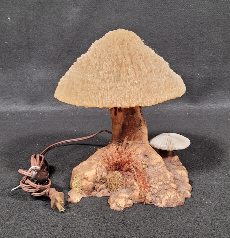 May include: A mushroom-shaped lamp with a textured, beige lampshade. The lamp's stem and base are brown, with a small mushroom and decorative foliage. The lamp has a brown cord and a two-prong plug.