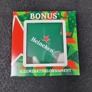 May include: A green and red Christmas-themed box with the word "BONUS" at the top. The box contains a green Heineken illuminating ornament. The ornament has the Heineken logo and the text "ILLUMINATING ORNAMENT".