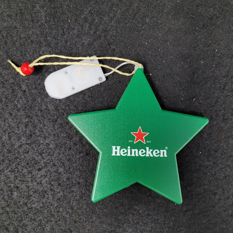 May include: A green star-shaped Heineken promotional item with a red star logo and white text. It has a small, white, rectangular light-up component attached by a string with a red bead.