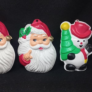May include: Three vintage Christmas decorations: a red-hatted Santa Claus with a white beard and a green holly sprig, a second Santa Claus ornament with a white beard and a green holly sprig, and a snowman decoration with a red hat and a green tree.