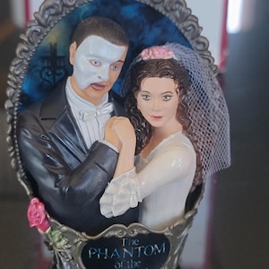 Può includere: Statuetta dei personaggi de Il Fantasma dell'Opera. Il personaggio maschile indossa una maschera e uno smoking, mentre il personaggio femminile indossa un abito bianco e un velo. La base recita "The Phantom of the Opera."