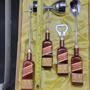 May include: A set of four miniature Johnnie Walker Scotch Whisky bottles with a bottle opener, a jigger, and a cocktail fork. The bottles are red and gold with the Johnnie Walker logo and text. The set is in a white box with a yellow lining.