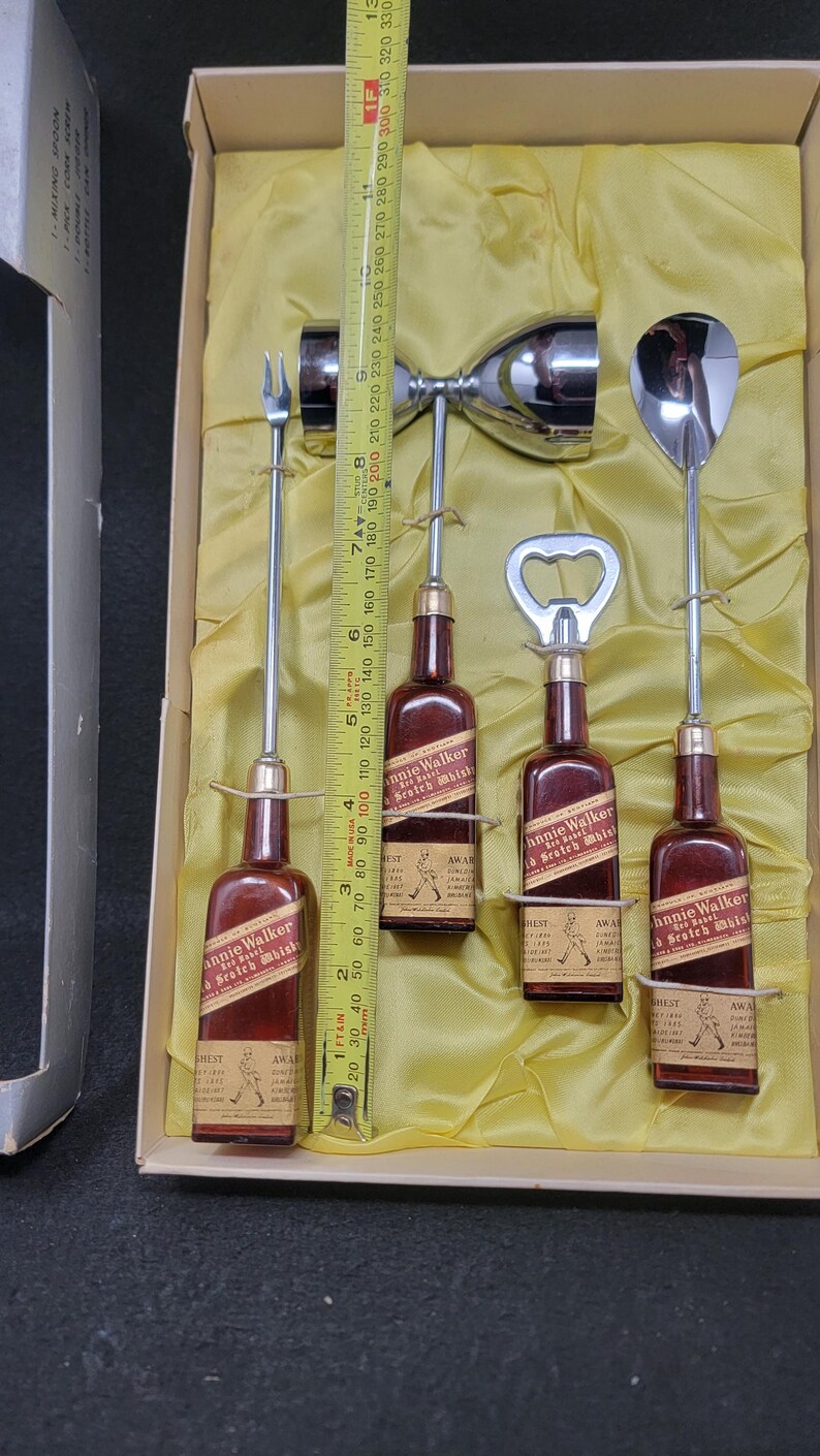May include: A set of four miniature Johnnie Walker Scotch Whisky bottles with a bottle opener, a jigger, and a cocktail fork. The bottles are red and gold with the Johnnie Walker logo and text. The set is in a white box with a yellow lining.