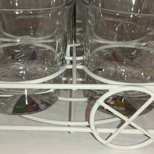 May include: White metal drink holder with four clear glass tumblers. The glasses have a decorative design etched into the glass.