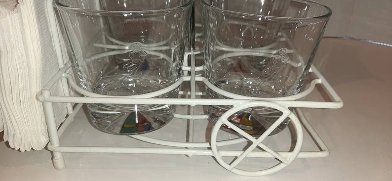 May include: White metal drink holder with four clear glass tumblers. The glasses have a decorative design etched into the glass.