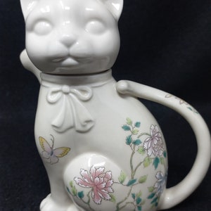 May include: White ceramic teapot shaped like a cat with floral designs and a butterfly. The teapot has a lid shaped like a cat's head.