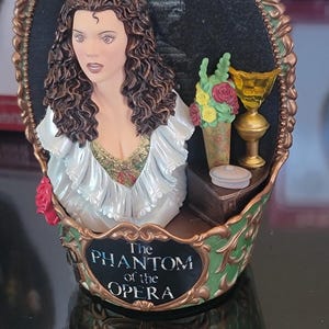 Può includere: Ornamento raffigurante un busto di donna con capelli castani ricci, che indossa un abito bianco. La base recita 'The Phantom of the Opera'. Include un calice dorato e un mazzo di fiori. La cornice è dorata e verde.
