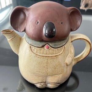 Może przedstawiać: Ceramiczny czajnik w kształcie misia koala. Głowa misia koala tworzy pokrywkę, z brązową twarzą i dużymi uszami. Korpus czajnika jest beżowy z teksturowanym wykończeniem, z małym uchwytem i wylewką.