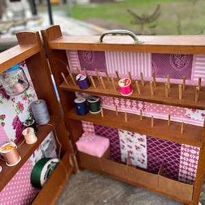 Op de afbeelding: Een houten naaidoos, open om planken en compartimenten te laten zien. De binnenkant is versierd met roze bloemen en patroonstof. Spoelen met draad in verschillende kleuren, een speldenkussen en andere naaispullen zijn zichtbaar.