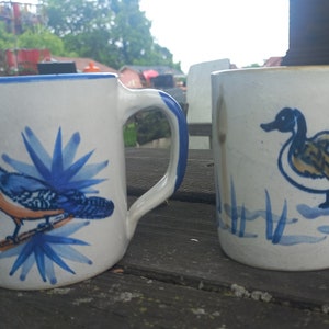 Könnte beinhalten: Zwei Keramikbecher mit blauen und weißen bemalten Designs. Ein Becher zeigt einen Blauhäher, der auf einem Ast mit blauen Blumen sitzt. Der andere Becher zeigt eine Ente, die in einem Teich mit blauen Schilf schwimmt.