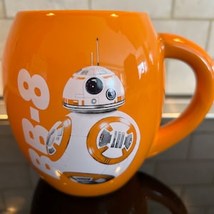 Może przedstawiać: Pomarańczowy kubek ceramiczny z nadrukiem białego i pomarańczowego bohatera Star Wars BB-8. Kubek ma zaokrąglony kształt i uchwyt.