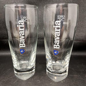 Peut inclure: Deux verres à bière transparents avec le mot "Bavaria" en blanc. Chaque verre présente un emblème bleu et or. Les verres sont vides et posés sur une surface sombre.