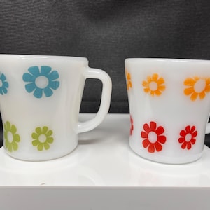May include: Two white ceramic mugs with floral patterns. One mug has blue and green flowers, the other orange and red. Both mugs have a curved shape and a white handle.
