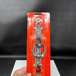 May include: A silver-toned metal door knocker with a vertical rectangular backplate featuring a textured center and decorative scrollwork at the top and bottom. The knocker is encased in clear plastic packaging on a red card with repeating text.