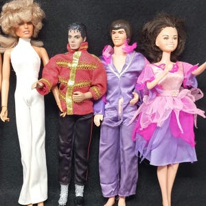 May include: A collection of vintage dolls, including a doll with blonde hair wearing a white jumpsuit, a doll with black hair wearing a red and gold jacket and black pants, a doll with brown hair wearing a purple jumpsuit, and a doll with brown hair wearing a pink and purple dress.