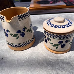 May include: A ceramic pitcher and a small jar with a lid. Both are cream-colored with a blue and green blueberry design and a black and white checkered border. The pitcher has a spout.