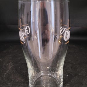 May include: A clear pint glass with a curved shape. The glass has the word "Guinness" in gold lettering on the side. The glass is empty and sits on a dark surface.