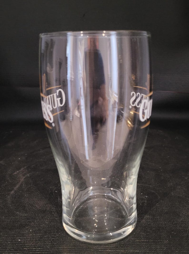 May include: A clear pint glass with a curved shape. The glass has the word "Guinness" in gold lettering on the side. The glass is empty and sits on a dark surface.