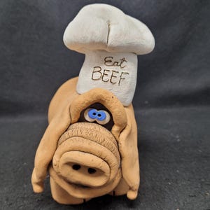 Può includere: Un maiale di ceramica marrone che indossa un cappello da chef bianco con le parole "Eat Beef" incise su di esso. Il maiale ha gli occhi blu e un grosso muso.
